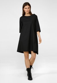 Svart A-linje klänning med båtkrage och trekvartsärmar. Slät tyg, mid-thigh längd och asymmetrisk fåll. Bärs med svarta snörstövlar.