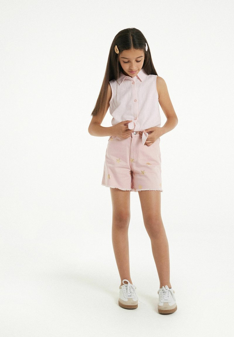 Růžová bezrukávová košile s límečkem, spárovaná s růžovými šortkami se zlatou hvězdnou výšivkou a roztrhanými lemy, doplněná bílými teniskami.