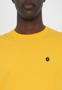 Sweatshirt galben din bumbac, cu guler rotund, având un mic logo hexagonal negru în zona stângă a pieptului. Textură netedă, design simplu.