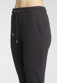 Schwarze Jogginghose mit elastischem Bündchen und Kordelzug; verfügt über Taschen und ein dezentes gesticktes Logo "H.I.S." am Bein.