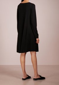 Schwarzes langärmliges Kleid mit rundem Halsausschnitt und einfacher Silhouette, das einen mittellangen Saum und eine weiche Textur aufweist. Getragen mit schwarzen Slippern.