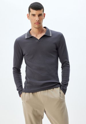 Grijze polo met lange mouwen en een V-hals kraag, voorzien van contrasterende witte strepen op de kraag, gemaakt van ribstof. Gecombineerd met beige broek.