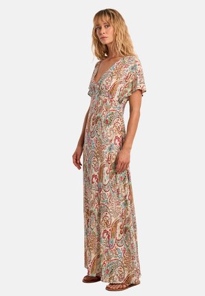 Donna in piedi che indossa un abito lungo multicolore con motivo paisley, maniche corte e sandali, su uno sfondo semplice.