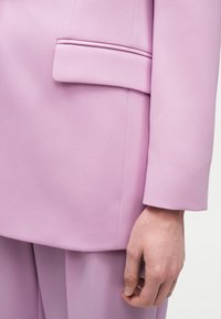 Κοντινό πλάνο ενός χεριού δίπλα σε ένα ραμμένο μωβ σακάκι με τσέπη με καπάκι και ταιριαστό μωβ παντελόνι.