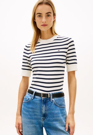 Vrouw draagt een wit met marineblauw gestreept shirt met korte mouwen en een klein logo, ingestopt in een blauwe spijkerbroek met een zwarte riem.