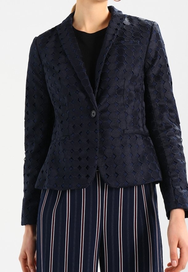 Femme portant un blazer texturé bleu foncé avec des motifs circulaires sur un top noir, associé à un pantalon bleu marine avec des rayures verticales blanches et rouges.
