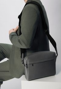 Siva kožna torba preko tijela s zaobljenim vrhom, teksturiranom površinom, zatvorenim džepom s patentnim zatvaračem i podesivim crnim remenom.