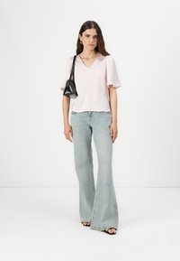 Blusa de cuello en V rosa claro con mangas cortas acampanadas, combinada con unos jeans acampanados azul claro y un bolso negro, sobre un fondo blanco.