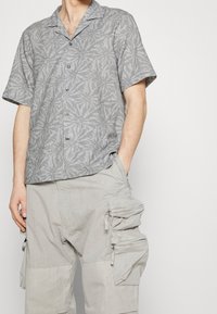 Szara koszula z krótkim rękawem z wzorem liści, zapinana na guziki z przodu, zestawiona z jasnoszarymi spodniami cargo z zamykanymi kieszeniami i teksturowaną tkaniną.