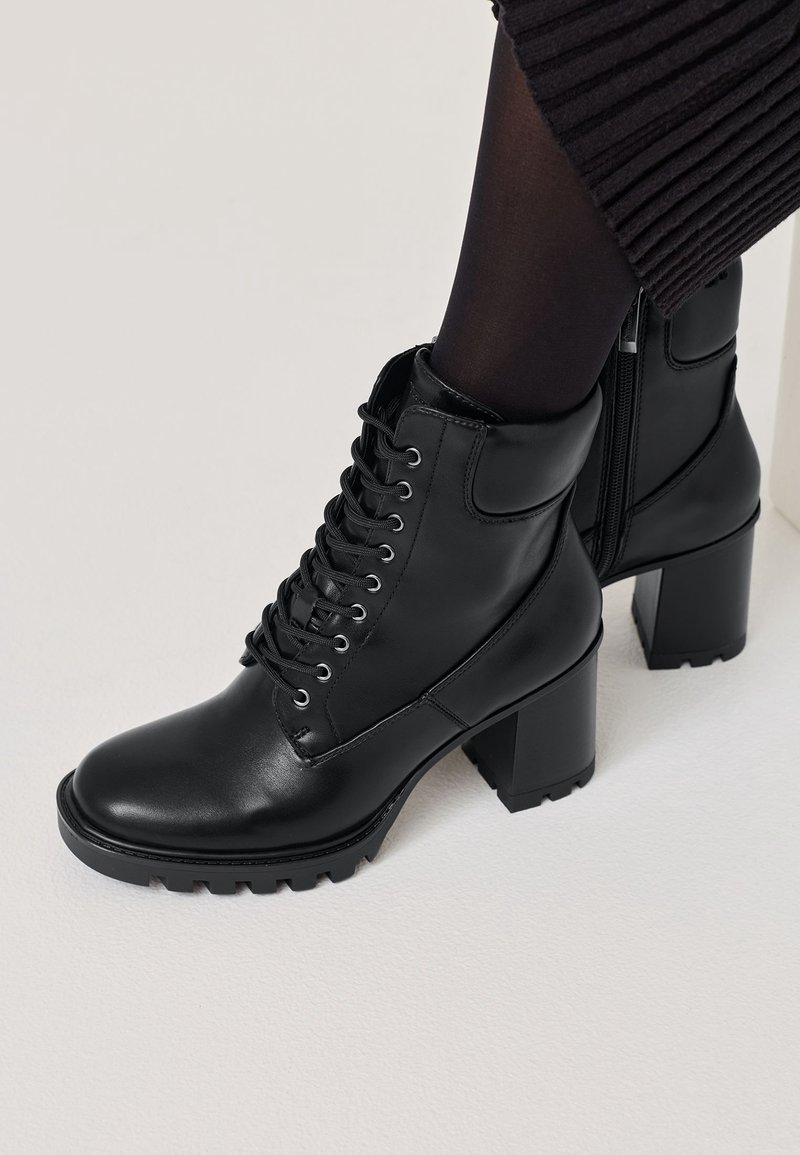 Bottes à lacets montantes en cuir noir avec talons blocs portées avec des collants noirs et une jupe ou un pantalon côtelé foncé sur un sol blanc.