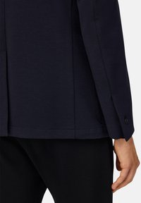 Blazer azul marino con un diseño estructurado, que presenta un puño de un solo botón, tela texturizada y un corte ajustado, combinado con pantalones negros.