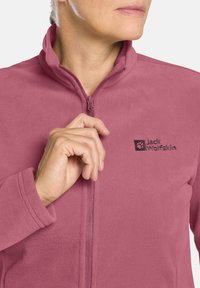 Giacca in pile di un rosa chiaro con colletto alto, zip frontale e logo ricamato sul lato sinistro. Tessuto morbido e testurizzato.
