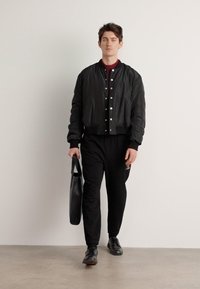 Zwarte bomberjack met drukknoopsluitingen, gedragen over een bordeaux shirt. Gecombineerd met zwarte broeken en schoenen, met een zwarte tote bag.