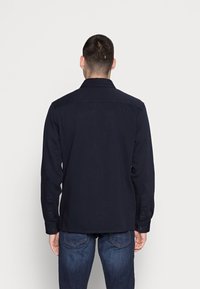 Chemise manches longues bleu marine avec col, poignets à boutons et texture unie. Portée avec un jean bleu foncé, vue de dos sur fond blanc.