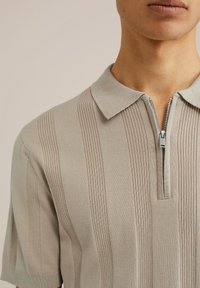 Camisa polo gris claro con cuello con cremallera, que presenta una textura de canalé vertical y mangas cortas. Diseño minimalista con un acabado suave.