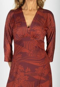 Kobieta w rudym kolorze sukienki z głębokim dekoltem w serek i rękawami do łokci, ozdobiona abstrakcyjnym wzorem liści oraz paskami.
