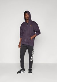 Casaco com capuz roxo com fecho de correr e um pequeno logótipo amarelo, combinado com calças de fato de treino pretas com riscas brancas nas laterais, usado com sapatilhas pretas.