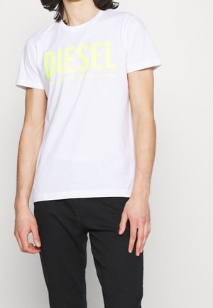 Homme portant un t-shirt blanc Diesel avec un logo jaune et un pantalon noir, debout devant un fond clair uni.