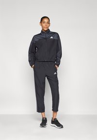Black athletic tracksuit with a zippered jacket and elastic waistband. Features gray side stripes and logo accents. Worn with black sneakers.