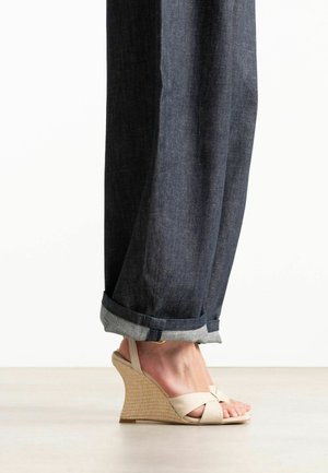 Persoon die opgerolde donkere spijkerbroek en beige sleehakken met geweven textuurhakken draagt op een lichte vloer.