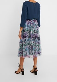 El vestido presenta una parte superior azul oscuro con un escote en forma de llave en la parte posterior, combinado con una falda transparente y fluida en un patrón de acuarela en tonos verde, morado y azul.