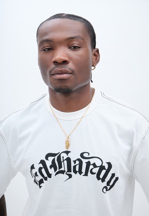 Bílá tričko s černým textem "Ed Hardy", s kontrastním šitím. Zlatý náhrdelník s listovým přívěskem doplňuje vzhled.