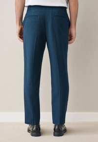 Man wearing blue tailored trousers and black dress shoes standing on beige carpet against a light wall.