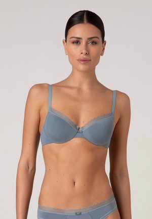 Donna con capelli scuri indossa reggiseno e mutandine azzurro chiaro abbinati con bordo trasparente, in piedi davanti a uno sfondo grigio chiaro uniforme.