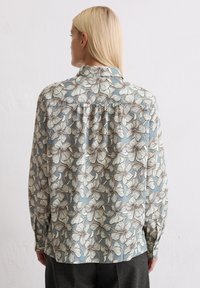 Long-sleeve blouse in light blue with white floral pattern and brown outlines. Features a collar and gathered back detail.