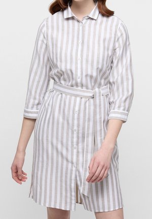 Kobieta ubrana w białą sukienkę z pionowymi beżowymi paskami, zapinaną na guziki, z dopasowanym paskiem z tego samego materiału i rękawami o długości do połowy przedramienia.