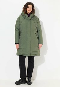Abrigo acolchado verde con capucha, que presenta un diseño aerodinámico, bolsillos laterales y una textura ligeramente brillante. Lleva puesto sobre pantalones negros y botas.