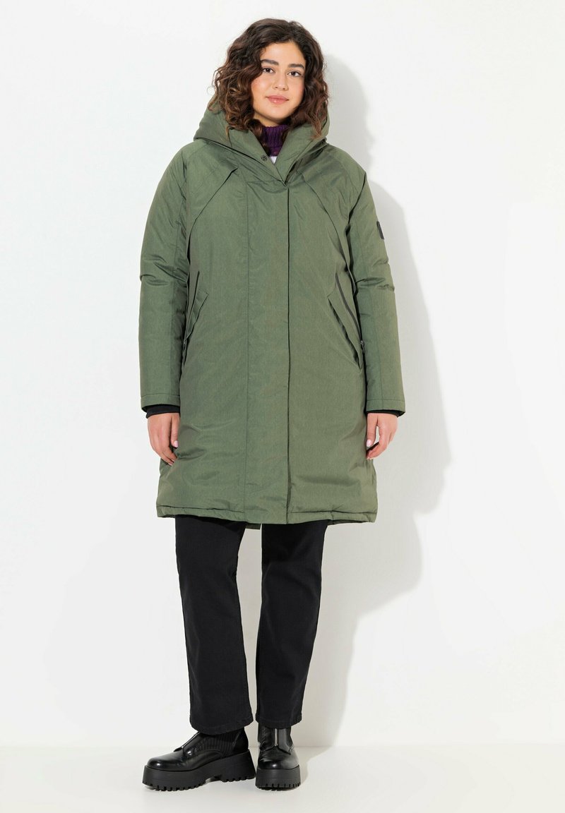 Abrigo acolchado verde con capucha, que presenta un diseño aerodinámico, bolsillos laterales y una textura ligeramente brillante. Lleva puesto sobre pantalones negros y botas.