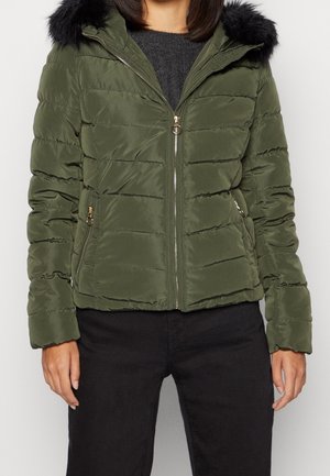 Femme portant une doudoune matelassée vert olive avec capuche en fausse fourrure noire, poches zippées et fermeture éclair dorée, associée à un pantalon noir.