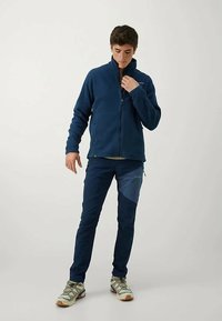 Giacca in pile blu marina con colletto alto e chiusura con zip, abbinata a pantaloni blu navy scuri con pannelli in tonalità più chiara. Modello che indossa scarpe da escursionismo grigie.