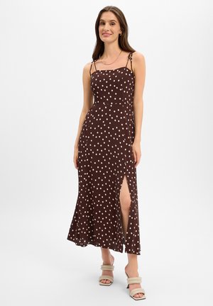 Frau trägt ein braunes ärmelloses Kleid mit Polka-Dots und seitlichem Schlitz sowie beigefarbene Sandalen mit Absatz, steht vor einem einfarbigen Hintergrund.