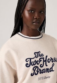 Beige sweatshirt met marineblauwe geborduurde tekst "The Two Horse Brand" en "1873 Levi's." Heeft een geribbelde ronde halslijn met contrasterende strepen.