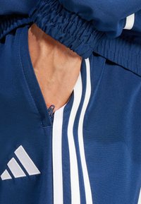 Chaqueta de cremallera navy con rayas blancas, puños elásticos y un logo bordado. Tela sintética suave con un área del cuello texturizada.