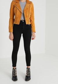 Suedefärgad senapsgul bikerjacka, grå crop top, svarta skinny jeans och svarta ankelskor med guldspännen. Slät textur, figursydd design.