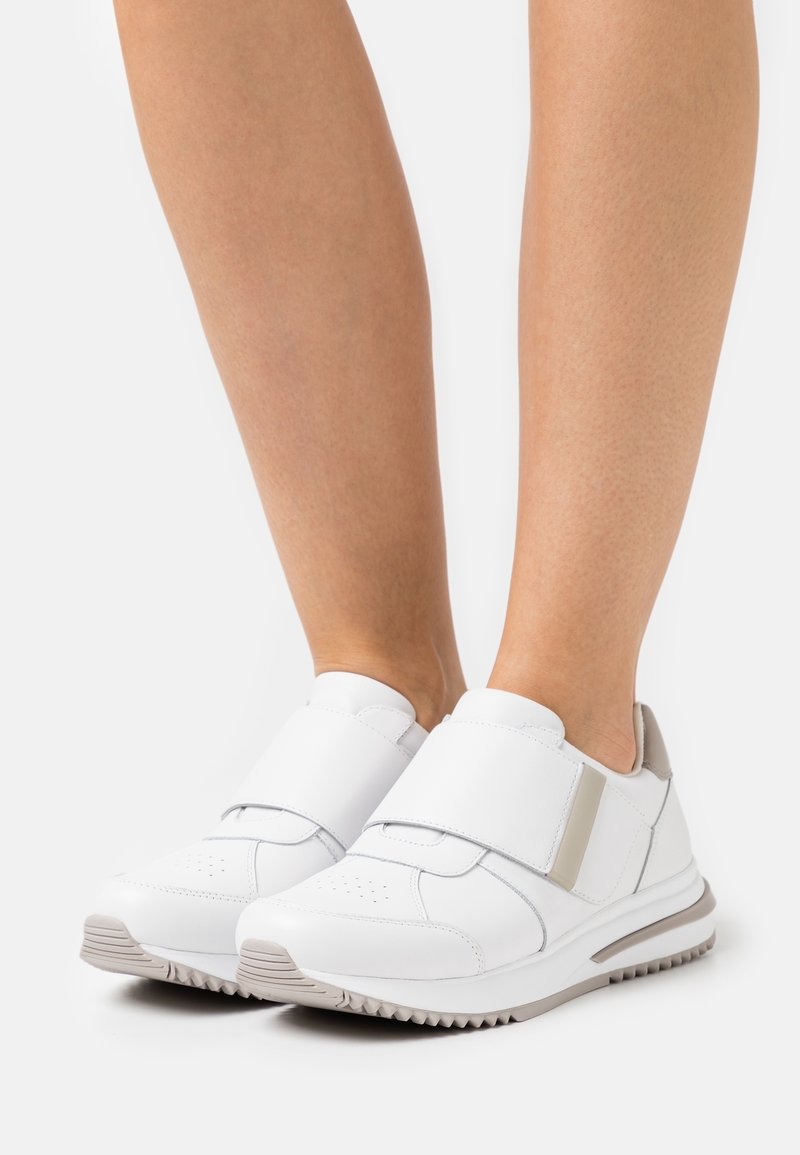 Baskets en cuir blanc avec larges bandes velcro portées sur des jambes nues, avec des accents beige et des semelles en caoutchouc texturé.