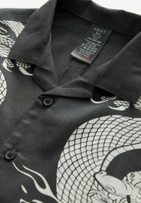 Svart kortärmad skjorta med en vit drakgrafik. Har en knäppt placket och en nedfällbar krage, tillverkad av texturerad tyg.