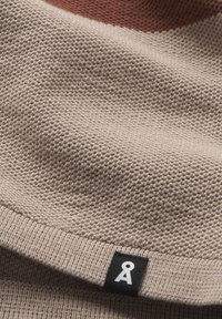 Indumento lavorato a maglia di colore beige con una superficie texture. Presenta un orlo a coste e un'etichetta nera con un logo bianco, parzialmente visibile.