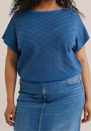 Femme portant un haut en maille bleu texturé à manches courtes, rentré dans une jupe en jean délavé moyen avec une couture avant et des poches.