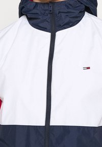 Marinblå och vit vindjacka med hög krage, framzipper och liten logotypdetalj på bröstet; slät textur och sportig design.