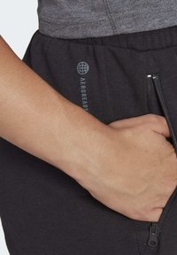 Pantalones de chándal negros con una textura suave, que cuentan con un bolsillo con cremallera y el logo "AEROREADY" en gris. Primer plano de la cintura y la mano.