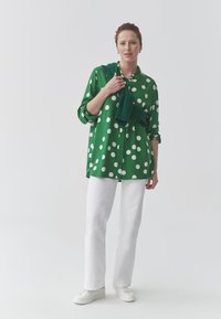 Žalia taškuota palaidinė su sagomis priekyje, balti kelnės ir balti sportbačiai. Žalias megztinis permestas per vieną petį. Paprastas dizainas.