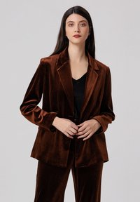 Blazer en velours marron avec revers à cran, manches longues et coupe décontractée. Associé à un haut noir, présentant une texture lisse et un brillant subtil.