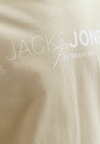 Μπεζ ύφασμα με λευκό τυπωμένο κείμενο «JACK & JONES Premium» σε διάφορα στυλ γραμματοσειράς στην επιφάνεια.
