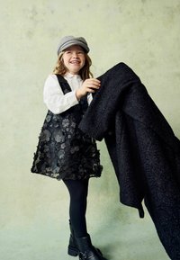 Mädchen mit grauem Hut, weißem T-Shirt, schwarzem Blumenkleid, schwarzen Strumpfhosen und Stiefeln, das einen großen schwarzen Mantel hält und vor einem schlichten Hintergrund lächelt.