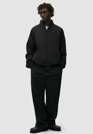 Uomo con dreadlocks corti che indossa una giacca nera con zip, pantaloni neri larghi e scarpe nere, in piedi con le mani nelle tasche.