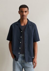 Donkerblauwe short-sleeve shirt van lichtgewicht stof met een kraag en knopen aan de voorkant; gedragen over een grijs T-shirt en lichte spijkerbroek.